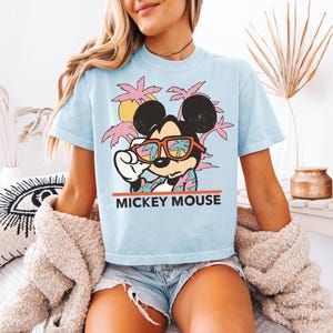 May include: Light blue t-shirt featuring a graphic of Mickey Mouse wearing sunglasses, with a tropical background of palm trees and a sunset. The text "MICKEY MOUSE" is printed in red below the graphic. The shirt is short-sleeved.