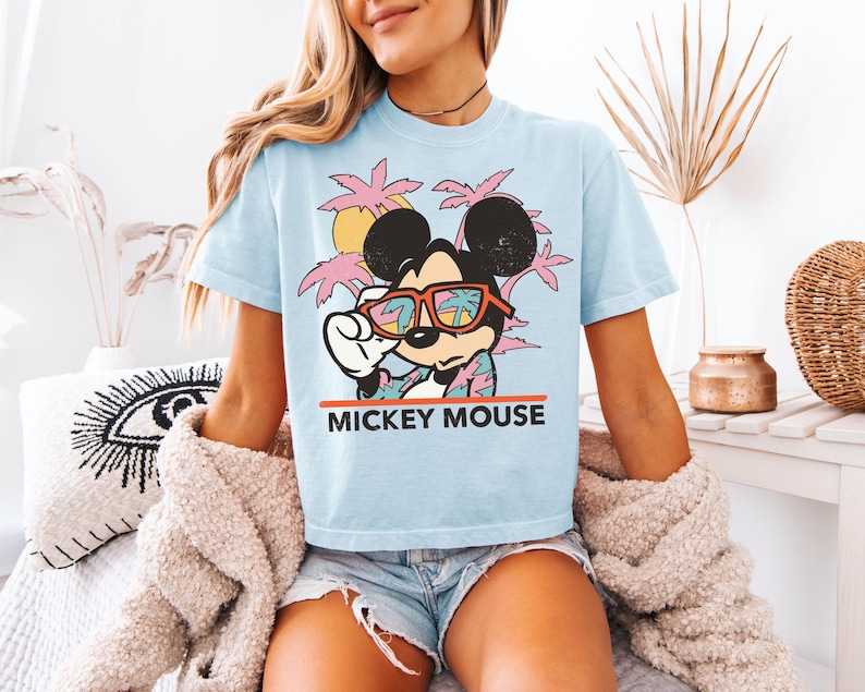 May include: Light blue t-shirt featuring a graphic of Mickey Mouse wearing sunglasses, with a tropical background of palm trees and a sunset. The text "MICKEY MOUSE" is printed in red below the graphic. The shirt is short-sleeved.