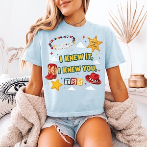 Może przedstawiać: Jasnoniebieski t-shirt z nadrukiem graficznym zawierającym tekst "I KNEW IT, I KNEW YOU,", bransoletkę z koralik&oacute;w z napisem "YOU'VE GOT A FRIEND IN ME,", gwiazdę szeryfa z napisem "INFINITY AND BEYOND,", czerwone kowbojki, czerwony kapelusz kowbojski oraz klocki "TS5".