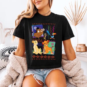 May include: Black t-shirt featuring a colorful graphic of characters from The Lion King. The design includes Simba, Timon, Pumbaa, and Zazu, set against a geometric background. The shirt is a casual wear item.