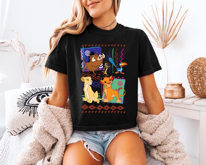 May include: Black t-shirt featuring a colorful graphic of characters from The Lion King. The design includes Simba, Timon, Pumbaa, and Zazu, set against a geometric background. The shirt is a casual wear item.