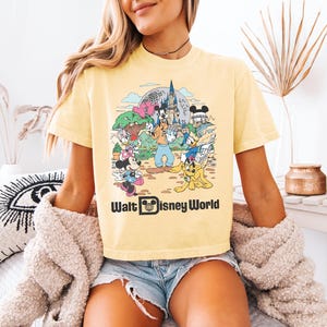 May include: Yellow t-shirt featuring a colorful graphic of Disney characters at Walt Disney World. The design includes Mickey Mouse, Minnie Mouse, Donald Duck, Goofy, and other characters in front of a castle and Spaceship Earth. The words "Walt Disney World" are printed below the graphic.