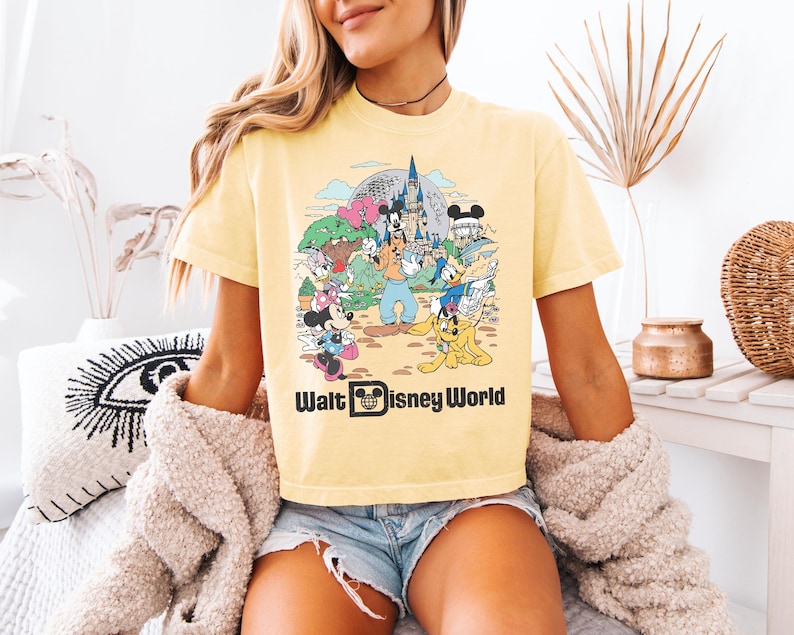 May include: Yellow t-shirt featuring a colorful graphic of Disney characters at Walt Disney World. The design includes Mickey Mouse, Minnie Mouse, Donald Duck, Goofy, and other characters in front of a castle and Spaceship Earth. The words "Walt Disney World" are printed below the graphic.
