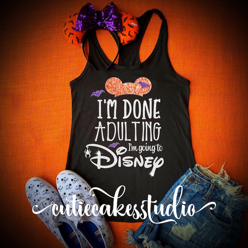 May include: Black tank top with glitter text that reads "I'm done adulting I'm going to Disney" with a Mickey Mouse head graphic. The tank top is on a orange background with a pair of black and white shoes and a pair of blue denim shorts.