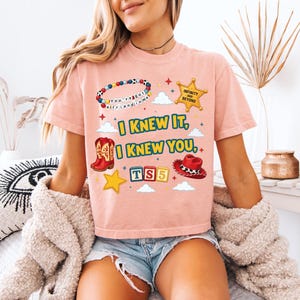 Może przedstawiać: Brzoskwiniowy t-shirt z nadrukiem graficznym przedstawiającym bransoletkę z koralik&oacute;w, złotą gwiazdę szeryfa z napisem "INFINITY AND BEYOND", czerwone kowbojki, czerwony kapelusz oraz klocki z napisem "TS5". Tekst "I KNEW IT, I KNEW YOU" jest wyśrodkowany.