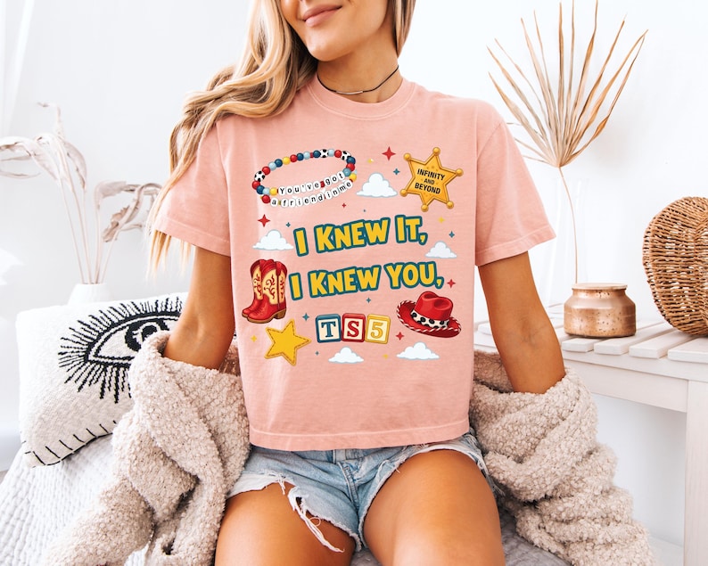 Może przedstawiać: Brzoskwiniowy t-shirt z nadrukiem graficznym przedstawiającym bransoletkę z koralik&oacute;w, złotą gwiazdę szeryfa z napisem "INFINITY AND BEYOND", czerwone kowbojki, czerwony kapelusz oraz klocki z napisem "TS5". Tekst "I KNEW IT, I KNEW YOU" jest wyśrodkowany.