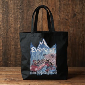 May include: Black tote bag featuring a colorful graphic of a roller coaster scene with cartoon characters, the word "Everest", and "Walt Disney World". The bag has two black handles and is made of a sturdy fabric.