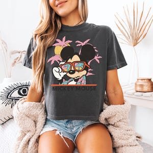 May include: Dark gray t-shirt featuring Mickey Mouse wearing sunglasses with a tropical design. The graphic includes pink palm trees, a yellow sun, and the text "MICKEY MOUSE" in white. The shirt is paired with denim shorts.