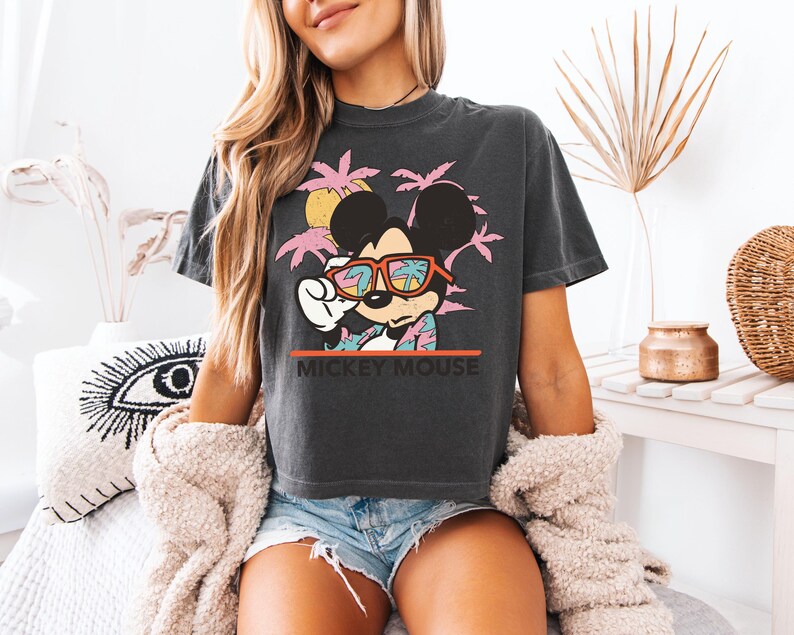 May include: Dark gray t-shirt featuring Mickey Mouse wearing sunglasses with a tropical design. The graphic includes pink palm trees, a yellow sun, and the text "MICKEY MOUSE" in white. The shirt is paired with denim shorts.