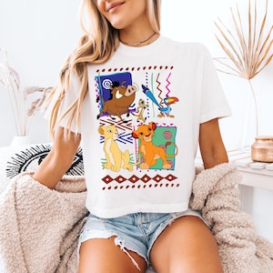 May include: White t-shirt featuring a colorful graphic of characters from The Lion King. The design includes Simba, Nala, Timon, Pumbaa, and Zazu, set against a geometric background with brown and blue accents.