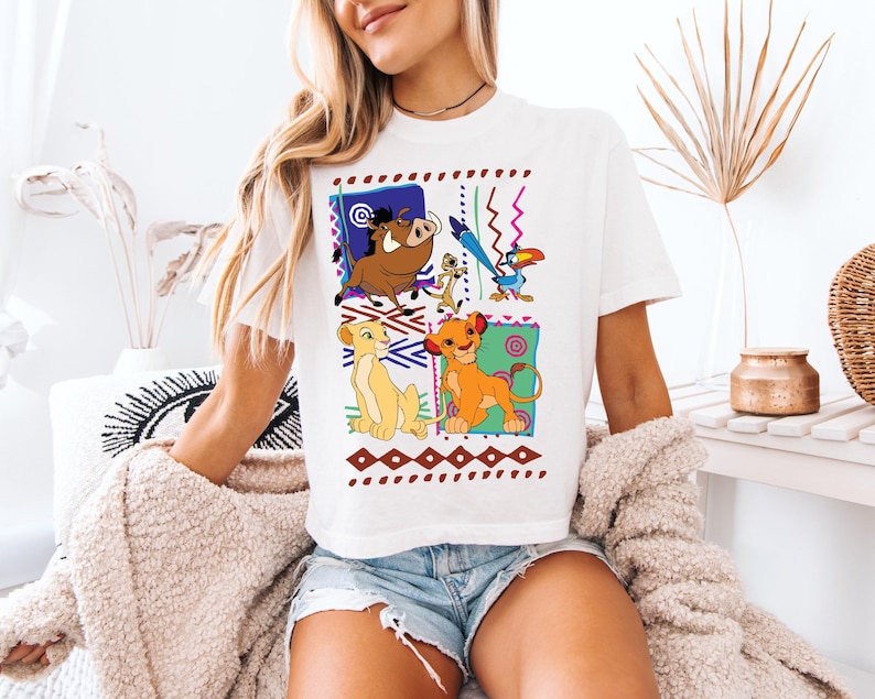 May include: White t-shirt featuring a colorful graphic of characters from The Lion King. The design includes Simba, Nala, Timon, Pumbaa, and Zazu, set against a geometric background with brown and blue accents.