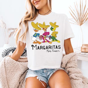 Op de afbeelding: Wit T-shirt met een cartoonontwerp van drie personages met sombrero's. De personages zijn in verschillende kleuren en poses. De tekst "Margaritas Por Favor" staat eronder in het zwart.