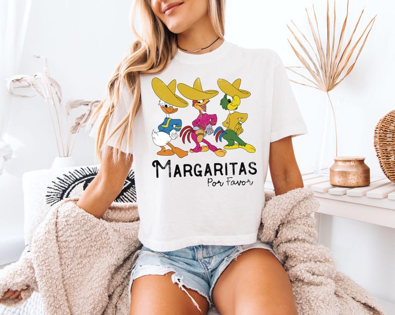 Op de afbeelding: Wit T-shirt met een cartoonontwerp van drie personages met sombrero's. De personages zijn in verschillende kleuren en poses. De tekst "Margaritas Por Favor" staat eronder in het zwart.