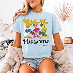 Op de afbeelding: Lichtblauw T-shirt met drie stripfiguren met sombrero's. De karakters zijn geel, blauw, roze en groen. De tekst "Margaritas Por Favor" staat eronder in het zwart gedrukt.