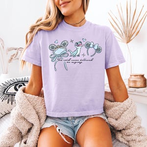 May include: A light purple t-shirt with a graphic featuring a blue bow, snowflake, and a cartoon lizard. The text "The cold never bothered me anyway" is printed on the shirt.