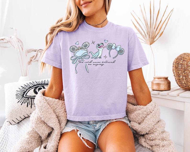 May include: A light purple t-shirt with a graphic featuring a blue bow, snowflake, and a cartoon lizard. The text "The cold never bothered me anyway" is printed on the shirt.