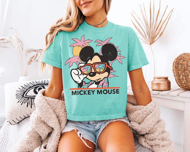 May include: A turquoise t-shirt featuring a graphic of Mickey Mouse wearing sunglasses, with a tropical sunset and palm trees. The text "MICKEY MOUSE" is printed in orange. The shirt is a relaxed fit.