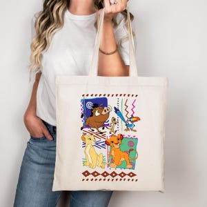 May include: A cream-colored tote bag featuring colorful illustrations of characters from The Lion King. The design includes Simba, Nala, Timon, Pumbaa, and Zazu, set against a backdrop of geometric patterns in blue, green, and brown.