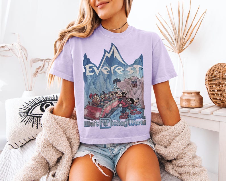 May include: A light purple t-shirt featuring a graphic of the Expedition Everest ride at Walt Disney World. The design includes the word "Everest" above a cartoon scene with Mickey Mouse and friends. The Walt Disney World logo is at the bottom.