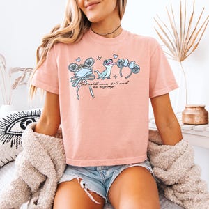 May include: A light pink t-shirt featuring a graphic design with the text "The cold never bothered me anyway." The design includes illustrations of a hat, a lizard, and mouse ears. The shirt is made of a soft material.