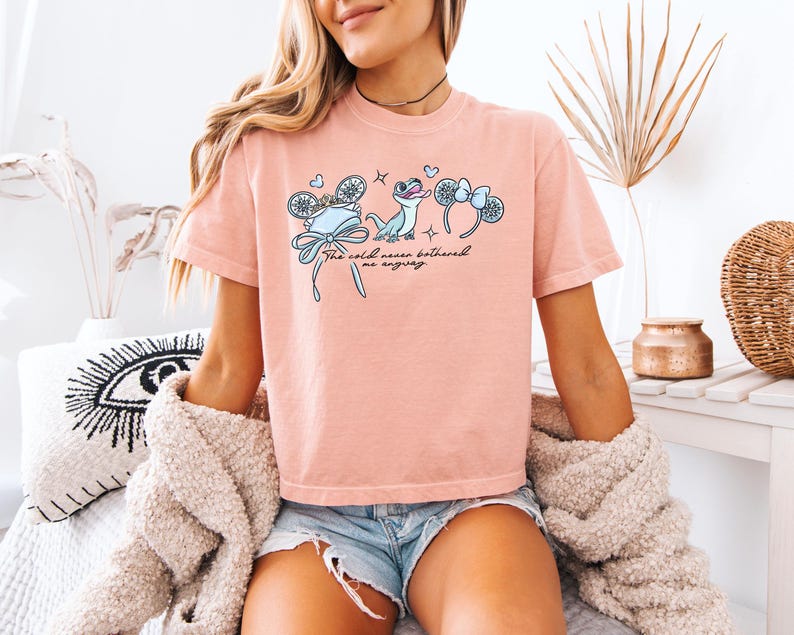 May include: A light pink t-shirt featuring a graphic design with the text "The cold never bothered me anyway." The design includes illustrations of a hat, a lizard, and mouse ears. The shirt is made of a soft material.