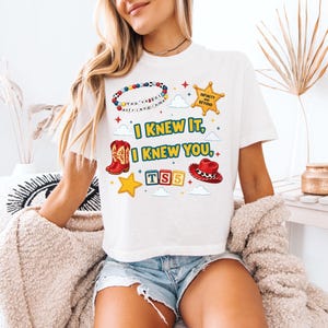 Może przedstawiać: Biały t-shirt z nadrukiem graficznym zawierającym tekst I KNEW IT, I KNEW YOU, TS5 oraz INFINITY AND BEYOND. Wz&oacute;r zawiera kolorową bransoletkę z koralik&oacute;w, ż&oacute;łtą gwiazdę szeryfa, czerwony kowbojski but, czerwony kowbojski kapelusz i ż&oacute;łte gwiazdki.