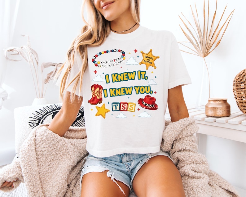 Może przedstawiać: Biały t-shirt z nadrukiem graficznym zawierającym tekst I KNEW IT, I KNEW YOU, TS5 oraz INFINITY AND BEYOND. Wz&oacute;r zawiera kolorową bransoletkę z koralik&oacute;w, ż&oacute;łtą gwiazdę szeryfa, czerwony kowbojski but, czerwony kowbojski kapelusz i ż&oacute;łte gwiazdki.