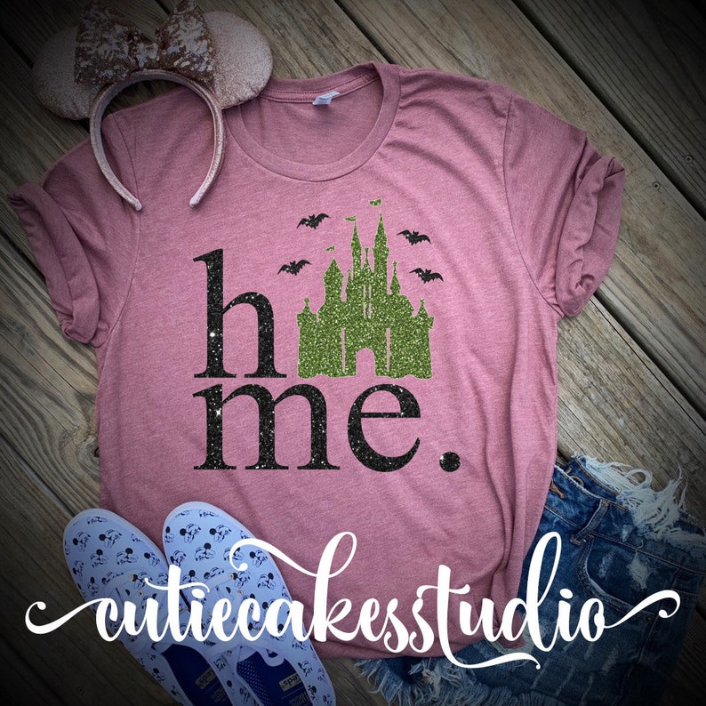 May include: A pink t-shirt with a green glitter castle graphic and the text "home" and "me" in black glitter. The castle has bats flying around it.