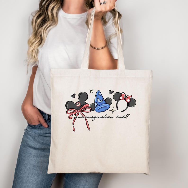 K&ouml;nnte beinhalten: Cremefarbene Tragetasche mit einem skurrilen Design mit Mickey Mouse-Ohren, einem blauen Zaubererhut und Minnie Mouse-Ohren. Das Design enth&auml;lt den Text "Some imagination. huh?" in einer verspielten Schriftart. Die Tasche wird von einer Person gehalten.