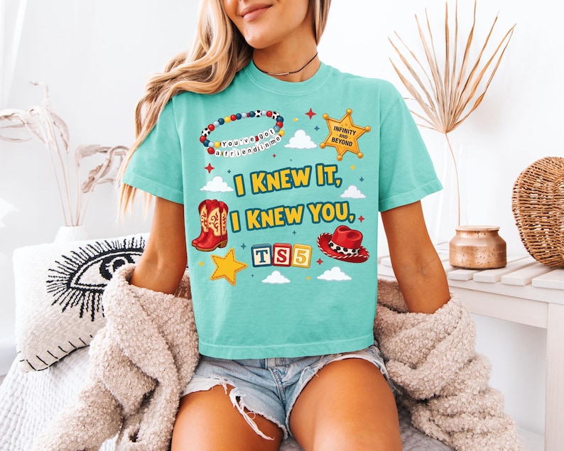 Może przedstawiać: Turkusowy t-shirt z nadrukiem graficznym zawierającym tekst "I KNEW IT, I KNEW YOU", bransoletkę z koralik&oacute;w z napisem "YOU'VE GOT A FRIEND IN ME", gwiazdę szeryfa z "INFINITY AND BEYOND", czerwone kowbojki, czerwony kapelusz oraz klocki z napisem "TS5".