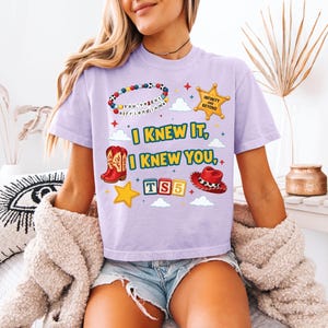 Może przedstawiać: Jasnofioletowy t-shirt z nadrukiem graficznym zawierającym tekst "I KNEW IT, I KNEW YOU,", bransoletkę z koralik&oacute;w, ż&oacute;łtą gwiazdę szeryfa, czerwone kowbojki, czerwony kapelusz kowbojski oraz kolorowe klocki z napisem "TS5" pośr&oacute;d chmur i gwiazdek.