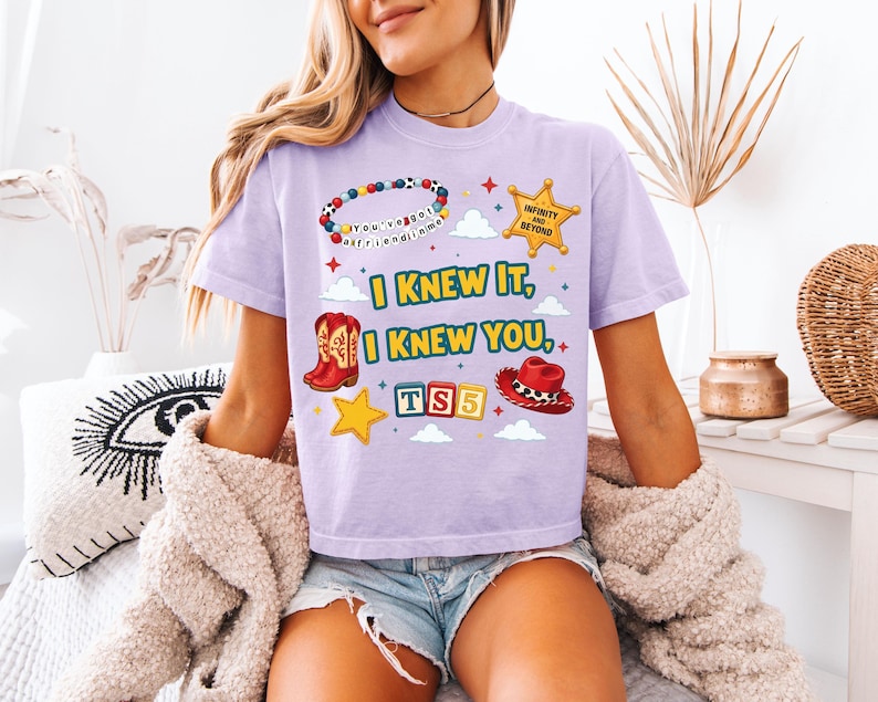 Może przedstawiać: Jasnofioletowy t-shirt z nadrukiem graficznym zawierającym tekst "I KNEW IT, I KNEW YOU,", bransoletkę z koralik&oacute;w, ż&oacute;łtą gwiazdę szeryfa, czerwone kowbojki, czerwony kapelusz kowbojski oraz kolorowe klocki z napisem "TS5" pośr&oacute;d chmur i gwiazdek.