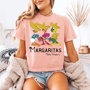 Op de afbeelding: Een lichtroze t-shirt met drie stripfiguren met sombrero's. De figuren zijn blauw, roze en geel. De tekst "Margaritas Por Favor" staat onder de figuren.