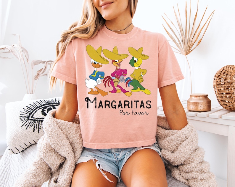 Op de afbeelding: Een lichtroze t-shirt met drie stripfiguren met sombrero's. De figuren zijn blauw, roze en geel. De tekst "Margaritas Por Favor" staat onder de figuren.