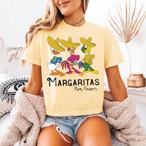 Op de afbeelding: Lichtgeel T-shirt met een cartoonontwerp van drie personages met sombrero's. De personages zijn blauw, paars en groen. De tekst "Margaritas Por Favor" staat eronder in het zwart.