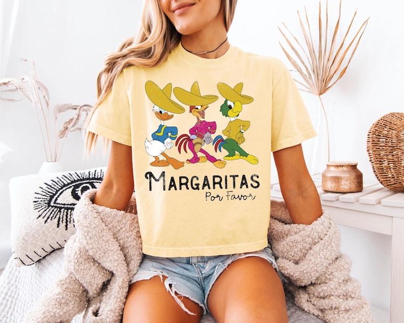 Op de afbeelding: Lichtgeel T-shirt met een cartoonontwerp van drie personages met sombrero's. De personages zijn blauw, paars en groen. De tekst "Margaritas Por Favor" staat eronder in het zwart.
