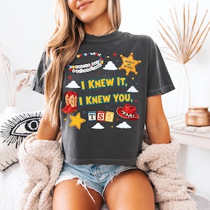 Może przedstawiać: Ciemnoszary t-shirt z kr&oacute;tkim rękawem z nadrukiem graficznym zawierającym tekst "I KNEW IT, I KNEW YOU," "YOU'VE GOT A FRIEND IN ME," "INFINITY AND BEYOND," oraz "TS5." Wz&oacute;r przedstawia kowbojski but, kapelusz, gwiazdę oraz chmury.