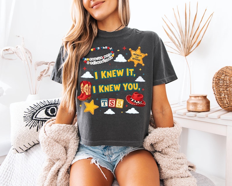 Może przedstawiać: Ciemnoszary t-shirt z kr&oacute;tkim rękawem z nadrukiem graficznym zawierającym tekst "I KNEW IT, I KNEW YOU," "YOU'VE GOT A FRIEND IN ME," "INFINITY AND BEYOND," oraz "TS5." Wz&oacute;r przedstawia kowbojski but, kapelusz, gwiazdę oraz chmury.