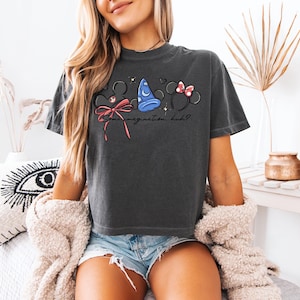 K&ouml;nnte beinhalten: Dunkelgraues T-Shirt mit einem skurrilen Design aus Mickey Mouse Ohren, einem blauen Zaubererhut und Minnie Mouse Ohren mit einer rosa Schleife. Der Text "Some imagination, huh?" ist darunter gedruckt.