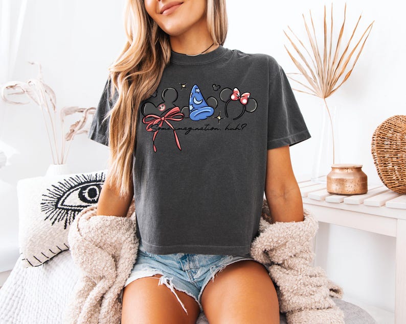 K&ouml;nnte beinhalten: Dunkelgraues T-Shirt mit einem skurrilen Design aus Mickey Mouse Ohren, einem blauen Zaubererhut und Minnie Mouse Ohren mit einer rosa Schleife. Der Text "Some imagination, huh?" ist darunter gedruckt.