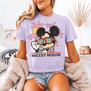 May include: A light purple t-shirt featuring a graphic of Mickey Mouse wearing sunglasses with a tropical scene reflected in the lenses. The text "MICKEY MOUSE" is printed in red below the graphic. The shirt is paired with denim shorts.