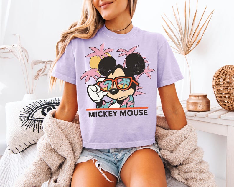 May include: A light purple t-shirt featuring a graphic of Mickey Mouse wearing sunglasses with a tropical scene reflected in the lenses. The text "MICKEY MOUSE" is printed in red below the graphic. The shirt is paired with denim shorts.