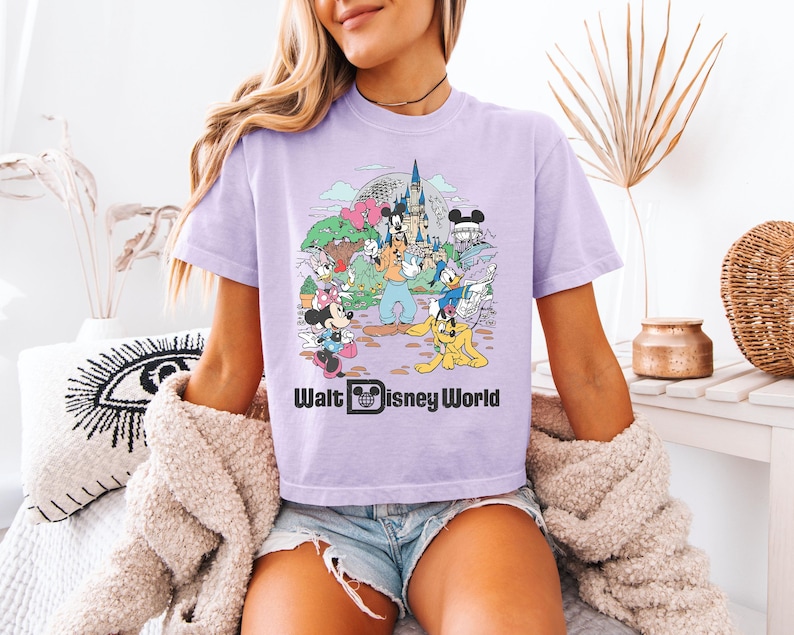 May include: A light purple t-shirt featuring a colorful graphic of Disney characters and landmarks, including Mickey Mouse, Minnie Mouse, and Cinderella's castle. The text "Walt Disney World" is printed below the graphic.