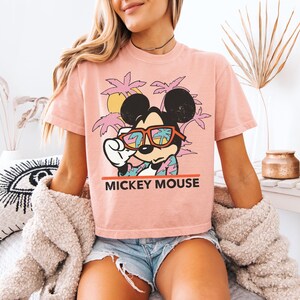 May include: A light pink t-shirt featuring Mickey Mouse wearing sunglasses with a tropical scene reflected in the lenses. The text "Mickey Mouse" is printed in red below the graphic. The shirt is paired with denim shorts.
