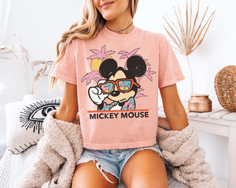 May include: A light pink t-shirt featuring Mickey Mouse wearing sunglasses with a tropical scene reflected in the lenses. The text "Mickey Mouse" is printed in red below the graphic. The shirt is paired with denim shorts.