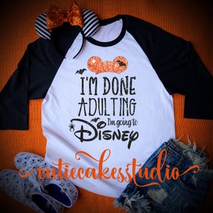 May include: A white and black baseball tee shirt with the text "I'm Done Adulting I'm going to Disney" in black and orange glitter. The shirt has a black and white striped headband with orange bows on top of it.