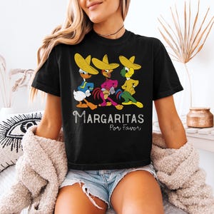 Op de afbeelding: Zwart T-shirt met drie stripfiguren met sombrero's. De tekst "Margaritas Por Favor" is wit. De karakters zijn geel, blauw, groen en roze.