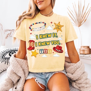 Może przedstawiać: Jasnoż&oacute;łty t-shirt z kr&oacute;tkim rękawem z nadrukiem graficznym zawierającym tekst "I KNEW IT, I KNEW YOU,", bransoletkę z koralik&oacute;w, gwiazdę szeryfa, czerwone kowbojki, czerwony kapelusz kowbojski oraz klocki z napisem "TS5" i małe gwiazdki.