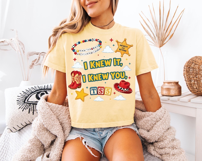 Może przedstawiać: Jasnoż&oacute;łty t-shirt z kr&oacute;tkim rękawem z nadrukiem graficznym zawierającym tekst "I KNEW IT, I KNEW YOU,", bransoletkę z koralik&oacute;w, gwiazdę szeryfa, czerwone kowbojki, czerwony kapelusz kowbojski oraz klocki z napisem "TS5" i małe gwiazdki.