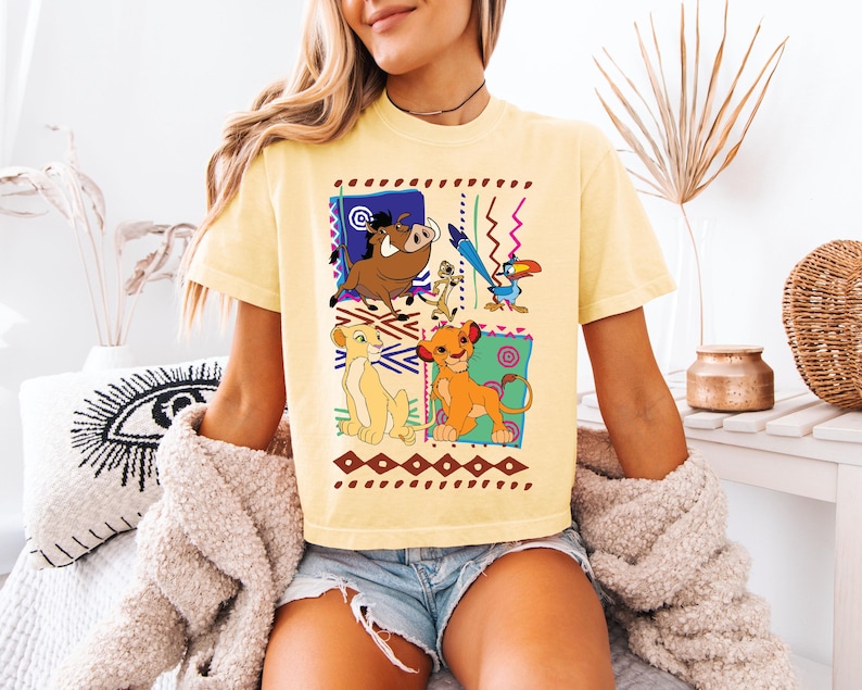 May include: Yellow t-shirt featuring a colorful graphic of characters from The Lion King. The design includes Simba, Nala, Timon, Pumbaa, and Zazu, set against a geometric background. The shirt is a casual, short-sleeved style.
